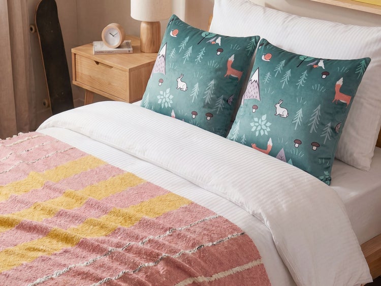 Zwei Zierkissen mit Waldtier-Motiv auf einem Bett mit weißer Bettwäsche und einer gestreiften Tagesdecke.