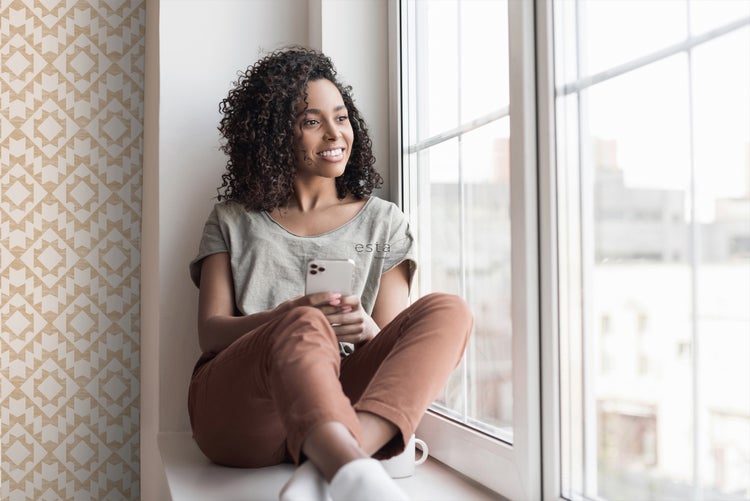 Frau sitzt mit einem Handy in der Fensterbank vor einer gemusterten Tapete