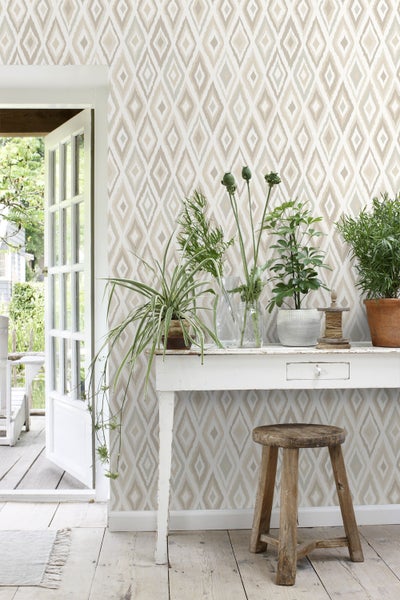 Raumansicht mit gemusterter Tapete, weißem Tisch, Pflanzen und Holzhocker