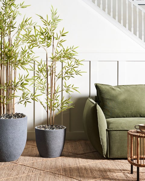 Innenraum mit zwei künstlichen Bambuspflanzen in strukturierten Pflanzgefäßen neben einem grünen Sofa