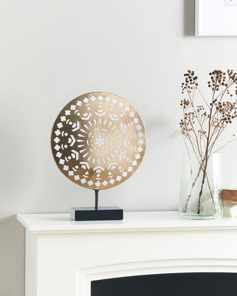 Dekorative goldfarbene Scheibe auf einem schwarzen Sockel als Tischdekoration neben einer Vase mit Trockenblumen.