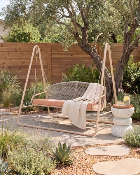 Gartenschaukel mit Geflecht und Sitzkissen auf einer Terrasse im mediterranen Stil mit Pflanzen und Holzzaun.