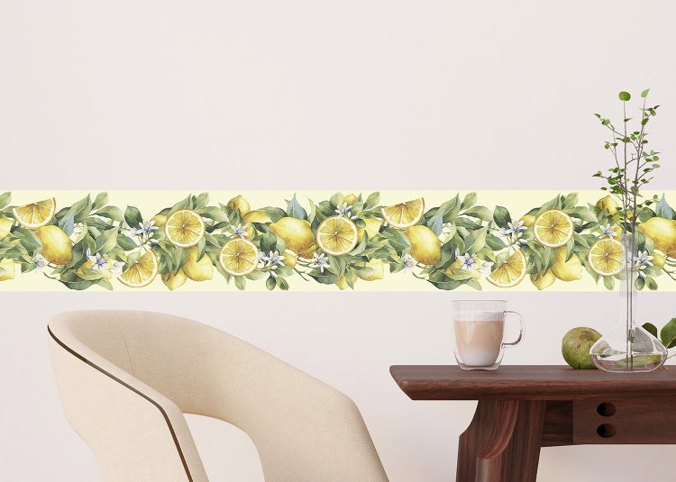 Tapetenbordüre mit Zitronenmotiv, Blättern und Blüten an einer Wand über einem Holztisch und einem hellen Sessel in heller Umgebung.