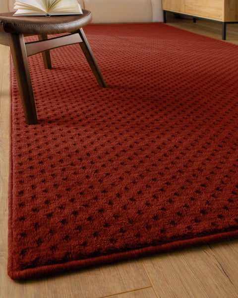Roter strukturierter Teppich mit Punktmuster auf Holzboden neben einem Hocker mit Buch.