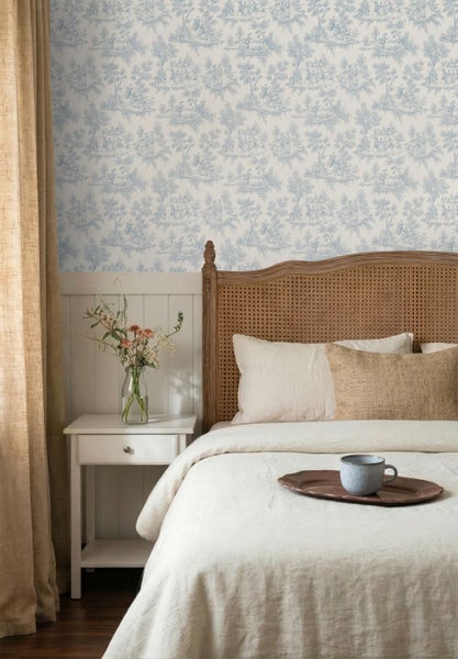 Schlafzimmer mit Toile-de-Jouy-Tapete mit blauen Landschaftsmotiven, Holzbett mit Rattan-Kopfteil, weißem Nachttisch und hellen Vorhängen.