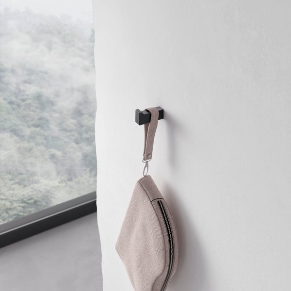 Ein moderner Wandhaken aus Metall, montiert an einer hellen Wand, an dem eine kleine Tasche mit Textilschlaufe hängt.