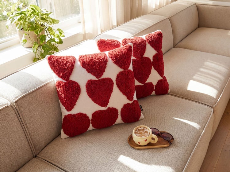 Zwei Dekokissen mit roten Herzmotiven auf einem hellen Sofa in einem hellen Wohnzimmer neben einer Pflanze.