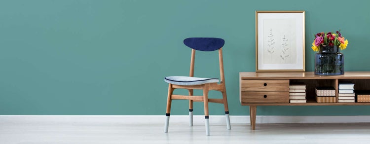 Modernes Wohnzimmer mit Holzstuhl, Sideboard aus Holz, Bilderrahmen und Blumen vor einer gestrichenen Wand auf hellem Boden.