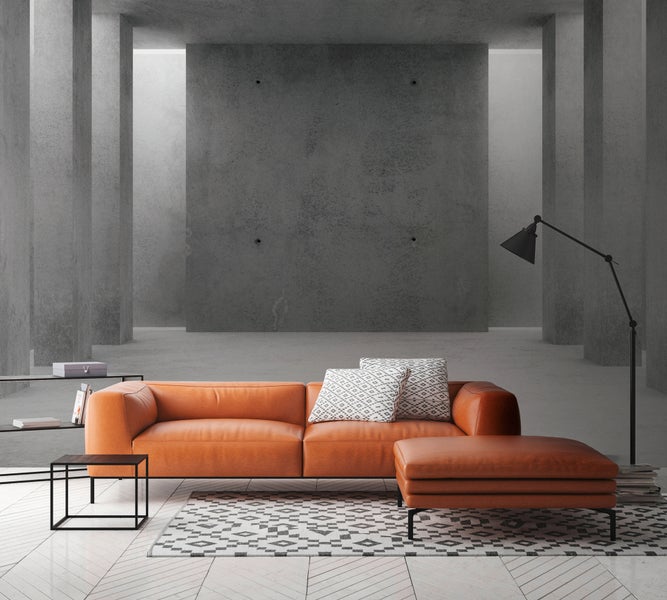 Modernes Wohnzimmer mit orangefarbenem Ledersofa, Ottoman, Betonwänden, Stehlampe und Teppich auf hellem Fischgrätparkett.