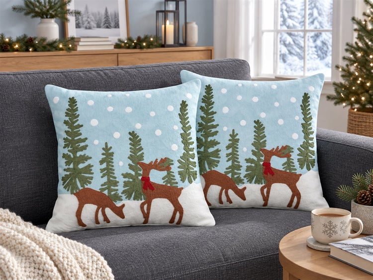 Zwei quadratische Dekokissen mit winterlichem Motiv aus Tannen und Rentieren auf einem Sofa in einem weihnachtlich dekorierten Wohnzimmer.
