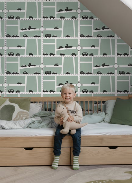 Kinderspielzimmer mit einer Mustertapete, die Straßen und Autos zeigt, einem Holzbett und einem lächelnden Kind mit Stofftier.
