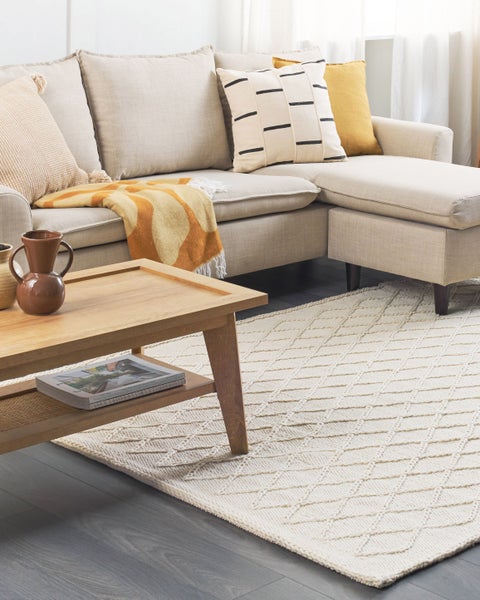 Helles Wohnzimmer mit hellem Ecksofa, dekorativen Kissen, gemusterter Decke, hölzernem Couchtisch und hellem Teppich mit Rautenmuster.