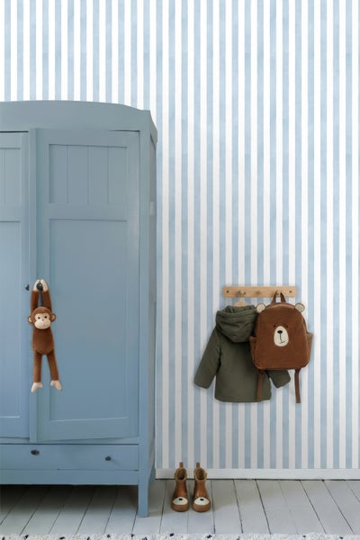 Tapete mit vertikalen Streifen in einem hellen Kinderzimmer mit blauem Kleiderschrank, hölzerner Hakenleiste und Bärenrucksack.