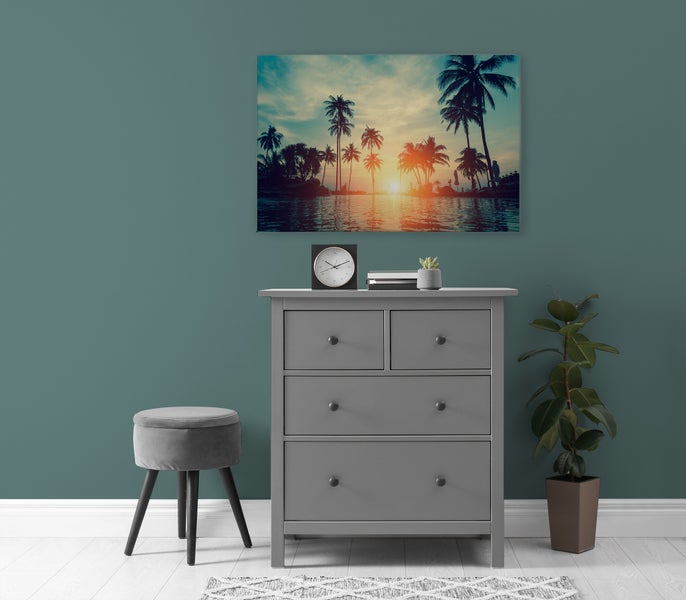 Graue Kommode mit vier Schubladen vor einer Wand mit Sonnenuntergangs-Wandbild, einem Hocker, einer Uhr und einer Zimmerpflanze im Wohnbereich.