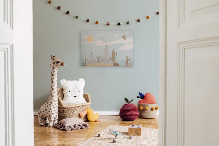 Ein Kinderzimmer mit einem Wandbild mit Wüstenmotiv, einem Giraffenkuscheltier, einem Rattan-Sessel, einem Teppich und Spielzeug auf Holzboden.