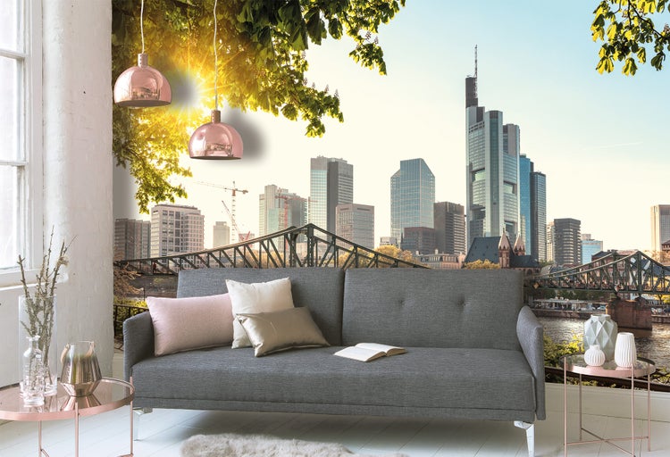 Fototapete mit der Skyline von Frankfurt, einer Brücke und Wolkenkratzern in einem Wohnzimmer hinter einem grauen Sofa.
