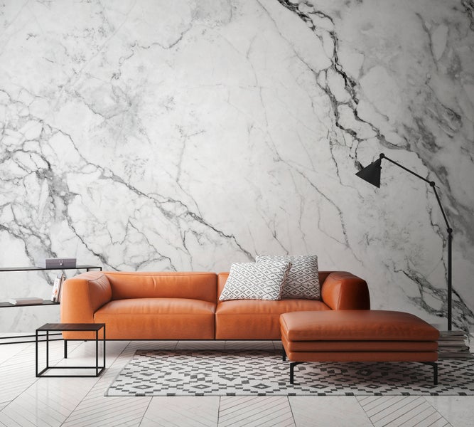 Heller Wohnraum mit Wandtapete in Marmor-Optik, orangefarbenem Sofa, Hocker, schwarzer Stehleuchte und geometrischem Teppich auf hellem Boden.