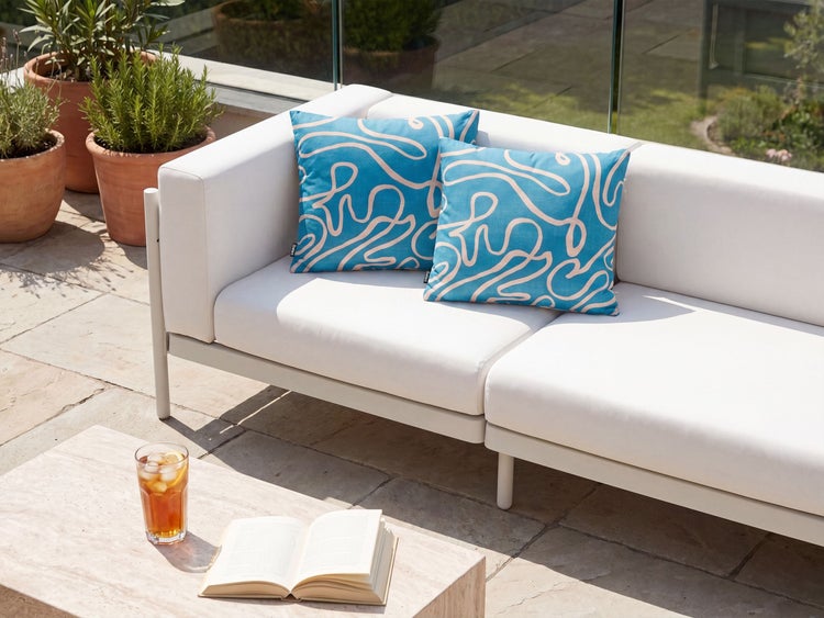 Zwei quadratische Kissen mit geschwungenem Linienmuster auf einem modernen Outdoor Sofa mit weißen Polstern auf einer Terrasse.