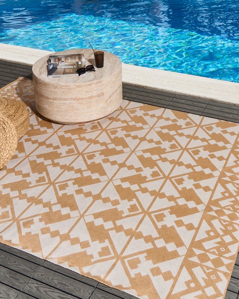 Outdoor-Teppich mit geometrischem Muster auf einer Terrasse am Pool, runder Beistelltisch in Steinoptik und geflochtene Sitzkissen.