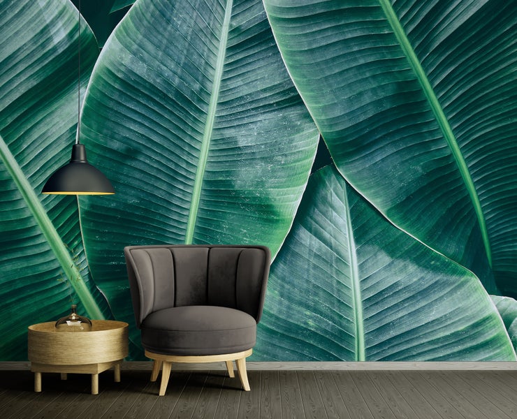 Modern gestalteter Raum mit großformatiger Fototapete mit grünen Bananenblättern, einem dunkelgrauen Sessel, einem Holztisch und einer schwarzen Lampe.