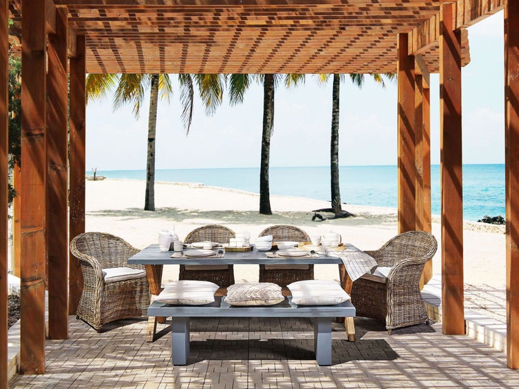 Gedeckter Esstisch mit Rattan-Stühlen und Bank unter einer Holzpergola am Strand