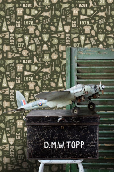 Dekorative Szene mit einem Spielzeugflugzeug auf einem beschrifteten Koffer vor einer Wand mit Sportmotiven