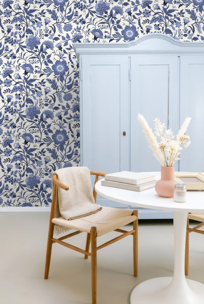 Vliestapete mit blauem Blumenmuster in hellem Zimmer mit hellblauem Schrank, Holzstuhl und weißem runden Tisch.