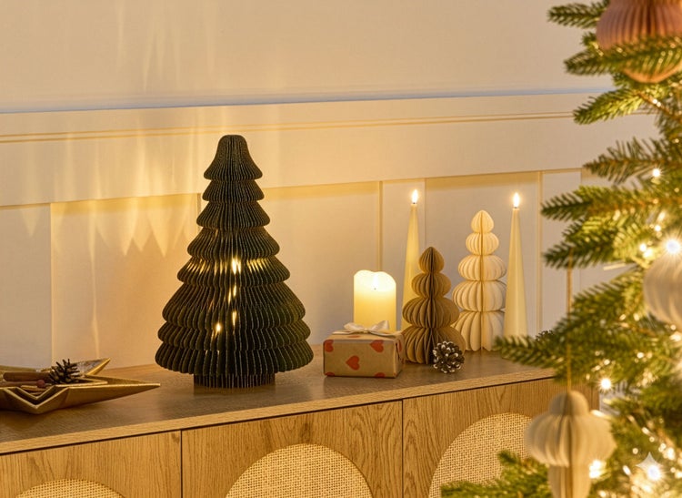 Festliche Dekoration mit beleuchtetem Papierweihnachtsbaum, Kerzen und Geschenk auf einer Kommode