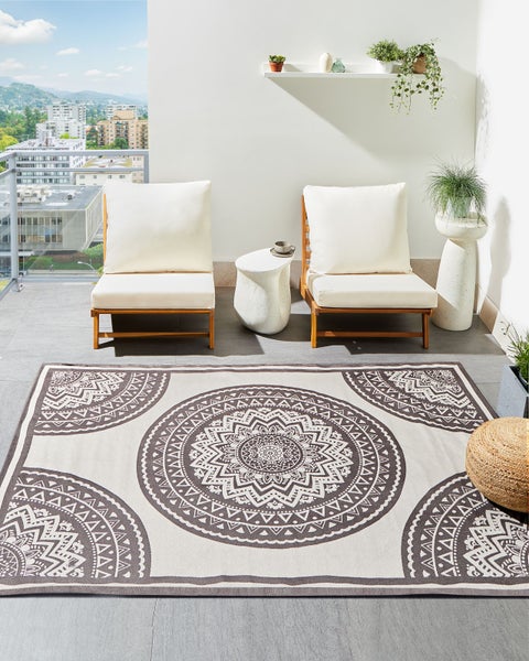 Heller Balkon mit zwei Holzsesseln, weißen Polstern, einem Beistelltisch und einem Outdoorteppich mit großem Mandala-Muster.