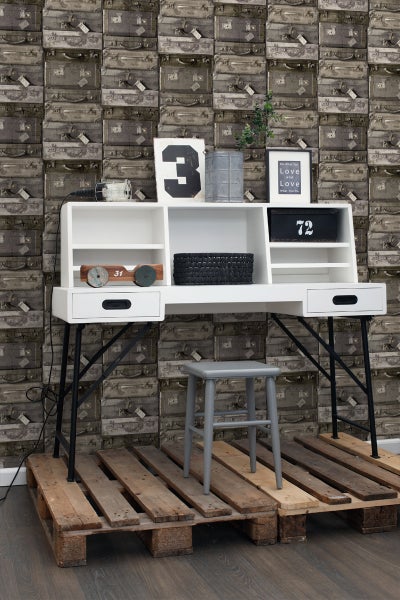 Schreibtisch mit Regalelementen, Stuhl und Dekoration auf einer Palette vor einer Tapete mit Koffermuster.