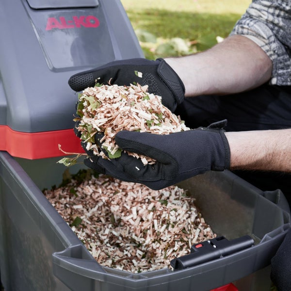 Hände in Arbeitshandschuhen halten frisch gehäckseltes Holzgut über dem Auffangbehälter eines AL-KO Gartenhäckslers.