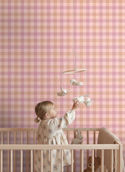 Kinderzimmer mit karierter Tapete, einem Holzgitterbett und einem Kind, das nach einem Mobile mit Stoffhasen greift.