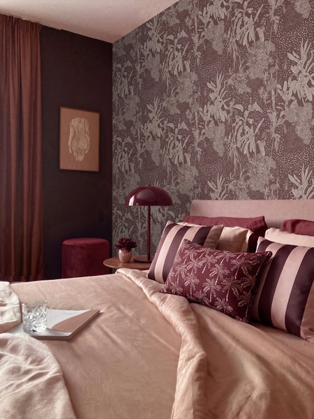 Schlafzimmer mit botanisch gemusterter Tapete, abgestimmter Bettwäsche, einer Tischleuchte in Pilzform und einem Samthocker.