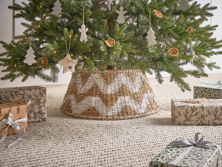 Weihnachtsbaum mit Baumschmuck, Lichterkette, geflochtenem Baumständer und verpackten Geschenken auf einem Teppich