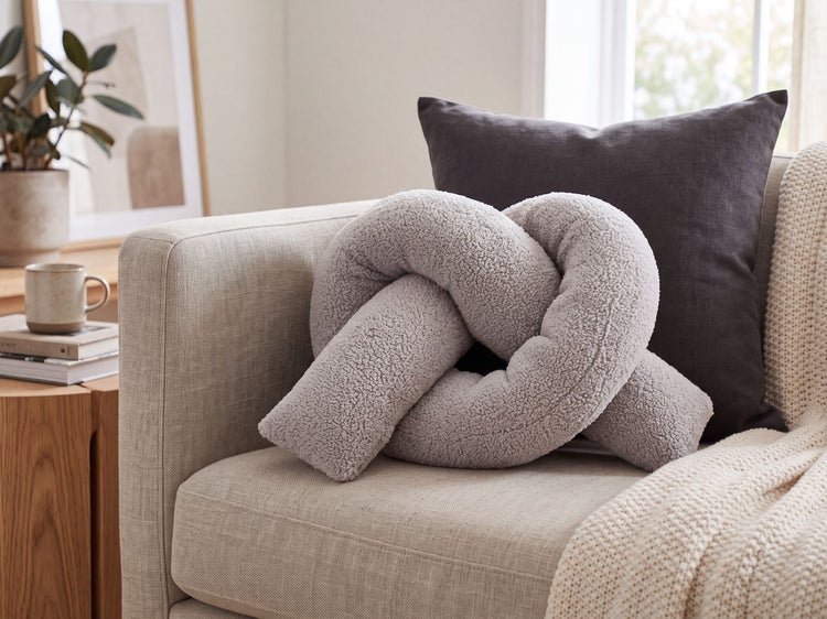 Graues Dekokissen in Knotenform aus strukturiertem Stoff auf einem Sofa platziert, ergänzt durch weitere Kissen und eine Wolldecke.