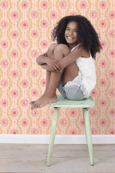 Ein Mädchen sitzt auf einem Hocker vor einer Wand mit Sonne Mond Tapete.