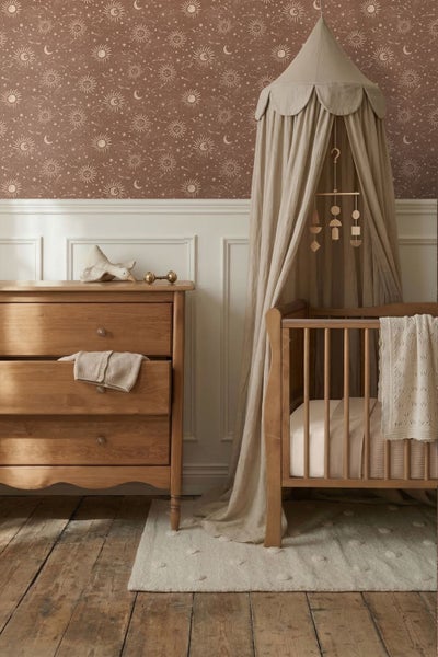 Kinderzimmer mit Tapete mit Sonnen- und Mondmuster, Holzkommode, Holzgitterbett mit Stoffbaldachin und weißer Wandvertäfelung.
