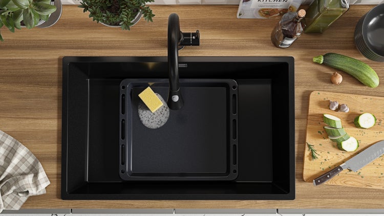 Schwarzes Küchenspülbecken mit Abtropfschale, Spülschwamm und Armatur auf einer Küchentheke mit Gemüse.