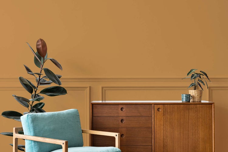 Wohnzimmer mit ockerfarbener Wandfarbe und Wandtäfelung, ausgestattet mit einem Holz-Sideboard, blauem Sessel und Zimmerpflanzen.