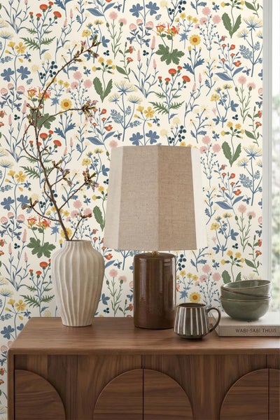 Vliestapete mit buntem Wiesenblumen-Muster hinter einer Holzkommode mit Vase, Tischlampe und Keramikgeschirr.