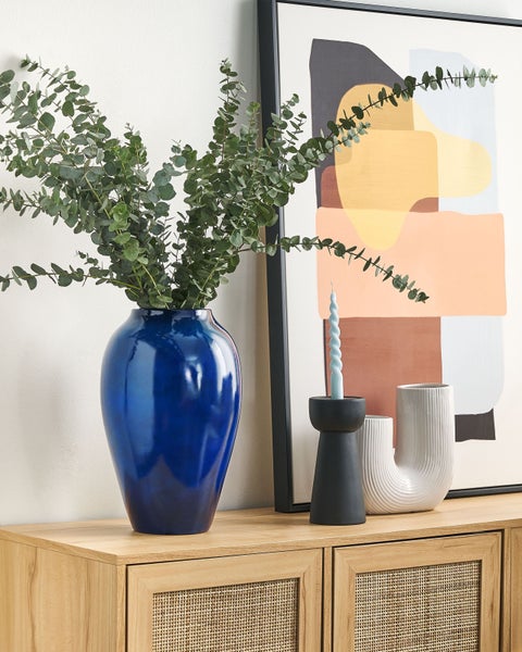 Dekoratives Arrangement mit blauer Vase, Eukalyptus, Kerzenhalter und abstraktem Bild auf einer Holzkommode.