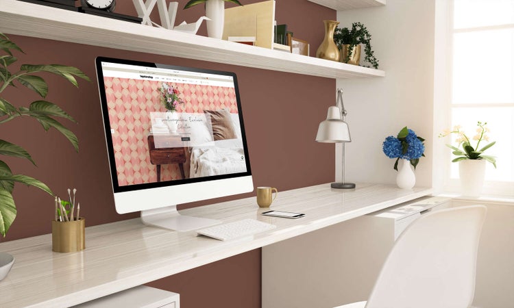 Hell eingerichteter Arbeitsplatz mit weißem Schreibtisch in Holzoptik, Monitor, Wandregalen und Dekoartikeln vor einer braun gestrichenen Wand.