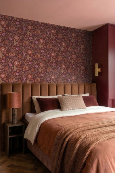 Ein Schlafzimmer mit floral gemusterter Tapete, einem gepolsterten Kopfteil am Bett, Bettwäsche, einer Strickdecke, Nachttisch und Tischleuchte.