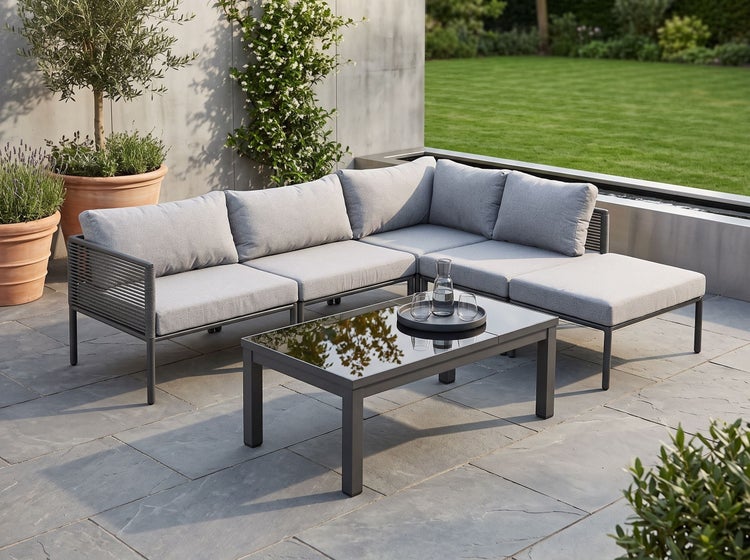 Moderne Loungegarnitur mit Ecksofa und Kaffeetisch auf einer Steinterrasse in einem Garten mit Pflanzen.