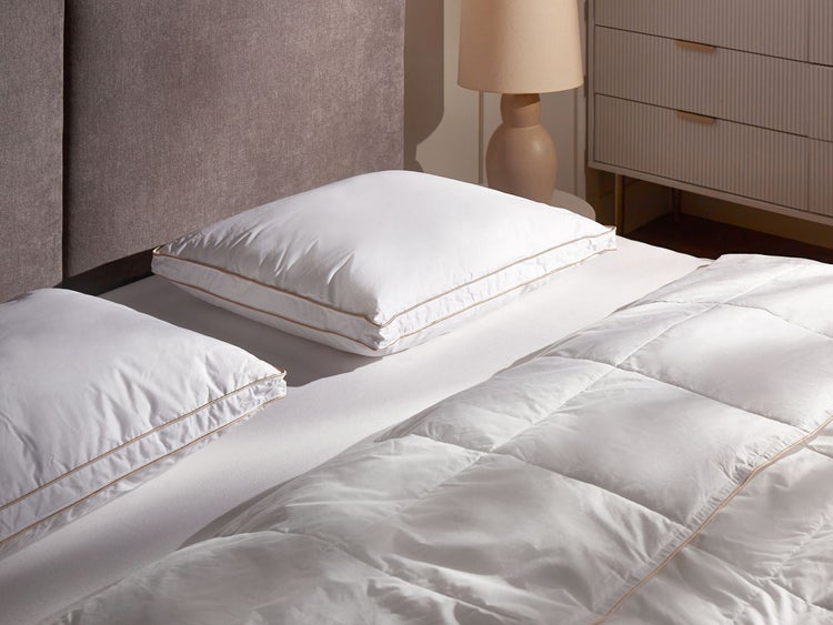 Helles Schlafzimmer mit zwei weißen Kopfkissen und gesteppter Bettdecke mit goldener Paspelierung auf einem Bett mit grauem Kopfteil.