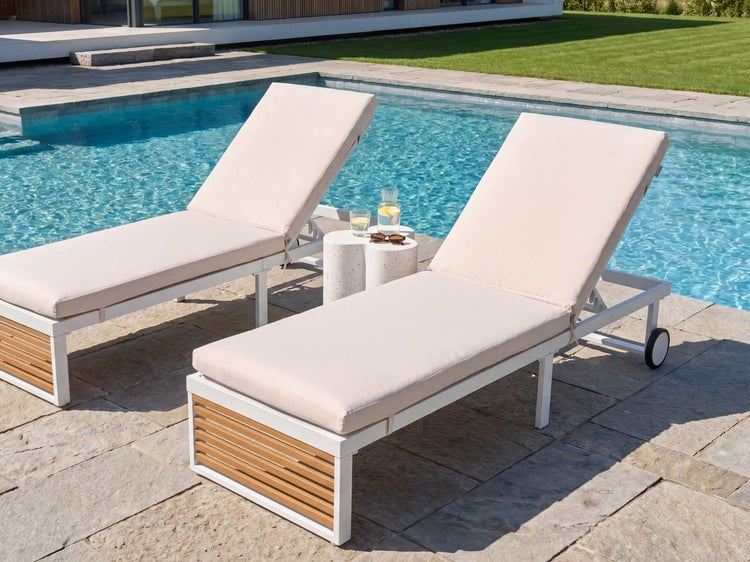 Zwei moderne Sonnenliegen mit weißem Metallgestell, Holzdetails und hellen Polstern auf einer Steinterrasse an einem Pool.
