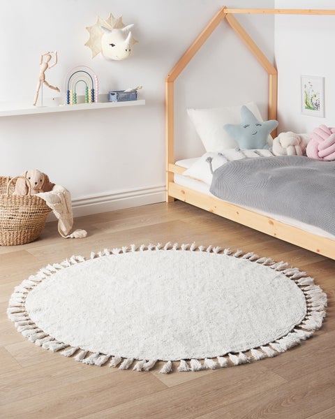 Helles Kinderzimmer mit rundem weißem Teppich mit Fransen auf Holzboden, einem Hausbett-Rahmen aus Holz und einem Wandregal mit Spielzeug.