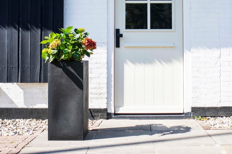 Hoher, rechteckiger Pflanzkübel in Schwarz mit Hortensie vor einer weißen Haustür auf einer gepflasterten Terrasse.