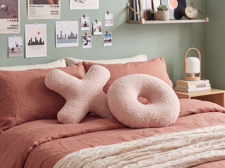 Gemütliches Schlafzimmer mit rosa Bettwäsche, zwei flauschigen Dekokissen in X- und O-Form sowie einem Nachttisch aus Holz mit Lampe.