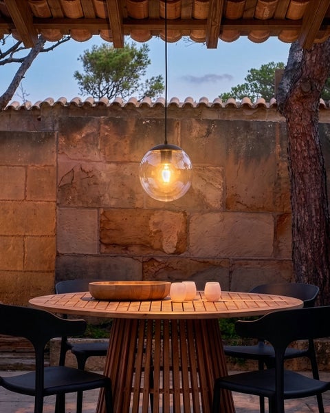 Moderne Pendelleuchte mit Glaskugel über einem runden Holztisch mit schwarzen Stühlen unter einem überdachten Terrassenbereich.
